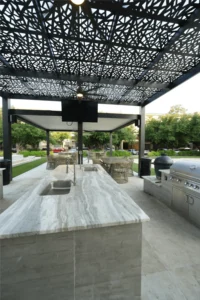 Metal pergola with geometric pattern over marble countertop, grill, bar stools, and mounted TV in landscaped yard